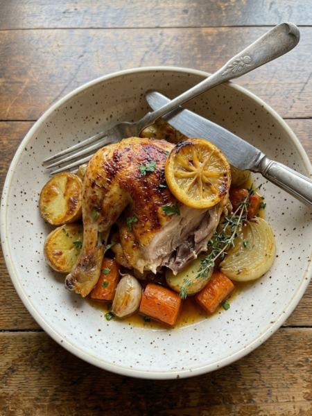 Roasted Cornish hen served with potatoes, carrots, onions, lemon slices, and herbs in a bowl.