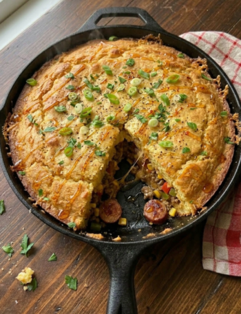 A rustic cast iron skillet filled with homemade Creamy Honey Butter Cornbread Cowboy Casserole baked until deeply golden brown. The casserole features a thick honey butter cornbread topping over a rich filling made with seasoned ground beef, smoky sausage, sweet corn, peppers, melted cheddar cheese, and creamy savory sauce finished with green onions, parsley, and hot honey drizzle.