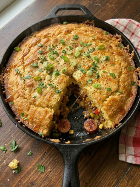A rustic cast iron skillet filled with homemade Creamy Honey Butter Cornbread Cowboy Casserole baked until deeply golden brown. The casserole features a thick honey butter cornbread topping over a rich filling made with seasoned ground beef, smoky sausage, sweet corn, peppers, melted cheddar cheese, and creamy savory sauce finished with green onions, parsley, and hot honey drizzle.